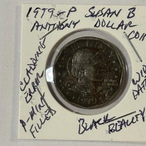Peut inclure: Une pièce d'un dollar Susan B. Anthony de 1979-P dans un support de protection. La pièce présente un profil de Susan B. Anthony et l'inscription "IN GOD WE TRUST". Des notes manuscrites sur le support incluent "CCADENCE ERROR" et "BLACK BEAUTY."