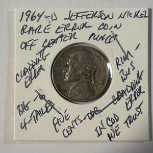 Op de afbeelding: Een 1964-D Jefferson nikkel munt met een zeldzame fout. De munt zit in een witte houder met handgeschreven aantekeningen die een excentrieke pons, bekledingsfout en andere details aangeven. De munt toont een profiel van Thomas Jefferson.