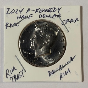 May include: A 2024 P-Kennedy half dollar coin, displayed on a white card. The coin features a profile of a person and the words "IN GOD WE TRUST." Handwritten notes on the card include "RARE," "ERROR," and "DOUBLING RIM."
