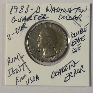 May include: A 1988-D Washington quarter coin in a protective holder. The coin is silver-colored, with the profile of George Washington visible. Handwritten notes on the holder indicate potential errors, including "Cladding Error" and "Double Edge Die."
