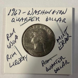 Może przedstawiać: Moneta Washington Quarter Dollar z 1967 roku, umieszczona w białym uchwycie. Ręcznie pisane notatki obejmują "Rim USDA", "Rim Liberty", "Mint Error" i "Rim Quarter Dollar". Moneta nosi ślady użytkowania.