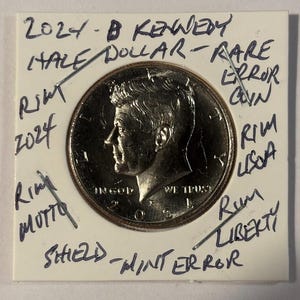 May include: A 2024 Kennedy half dollar coin, with the profile of John F. Kennedy. The coin is labeled with handwritten notes: "Rare Error Coin", "Mint Error", and other annotations. The coin is in a protective holder.