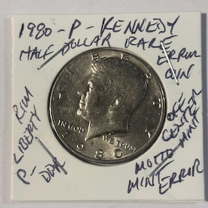 Puede incluir: Una moneda de medio dólar Kennedy de 1980-P, etiquetada como rara, con notas manuscritas que indican errores. La moneda está en un soporte protector, con la inscripción "IN GOD WE TRUST" visible.