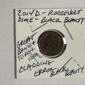 May include: A 2014-D Roosevelt dime, labeled "Black Beauty," encased in a protective holder. Handwritten text includes "Great Band + Total Cladding Error Black Beauty."