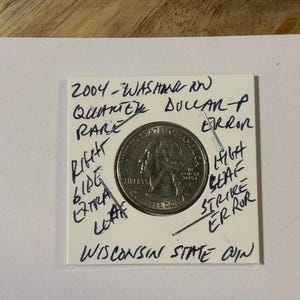 May include: A silver quarter coin from 2004, labeled as a Wisconsin State coin with a rare error. The coin is mounted on a white card with handwritten notes indicating a high leaf strike error and other details.