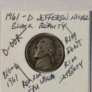 Może przedstawiać: Nikiel Jefferson z 1961-D, oznaczony jako "Black Beauty", jest prezentowany w ochronnym uchwycie. Moneta przedstawia profil Thomasa Jeffersona oraz napisy "In God We Trust" i "Liberty". Ręcznie pisane notatki otaczają monetę.