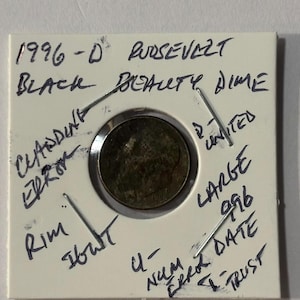 May include: A 1996-D Roosevelt dime, described as a "Black Beauty Dime," presented in a white holder. Handwritten annotations mention "Cladding Error," "Rim," and other details. The coin has a dark, aged appearance.