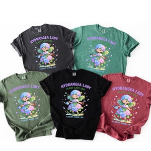 Puede incluir: Cinco camisetas en varios colores: negro, gris oscuro, verde, rojo y verde oliva. Cada camiseta presenta el texto "HYDRANGEA LADY PLANT LADY ERA" y una ilustración de dibujos animados de una mujer rodeada de hortensias. También está impresa la frase "FLORIST MODE: ON".