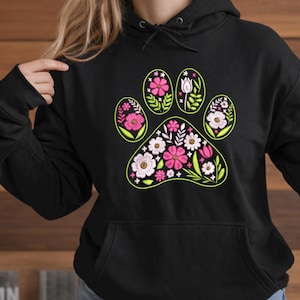 Könnte beinhalten: Schwarzer Kapuzenpullover mit einem floralen Pfotenabdruck-Design. Der Pfotenabdruck ist neongrün umrandet und mit rosa und weißen Blumen sowie grünen Blättern gefüllt. Der Pullover hat eine Vordertasche und eine Kapuze mit Kordelzug.