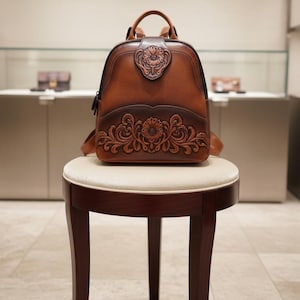 May include: A brown leather backpack featuring intricate floral embossing. The bag has a top handle, adjustable straps, and a front flap with a floral design. It rests on a small stool with a cream cushion. The bag's dimensions are approximately 30 cm in height.