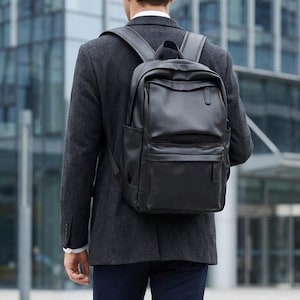 Könnte beinhalten: Ein schwarzer Lederrucksack mit mehreren Fächern und verstellbaren Trägern. Der Rucksack wird über den Schultern einer Person getragen, die einen dunkelgrauen Blazer und eine marineblaue Hose trägt. Im Hintergrund ein modernes Gebäude.