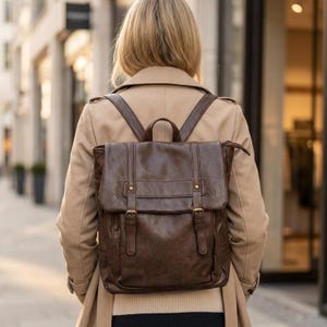 Może przedstawiać: Brązowy skórzany plecak z klapą, detalami w postaci klamer i regulowanymi paskami. Plecak jest noszony na plecach osoby ubranej w beżowy płaszcz.