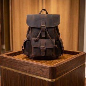 Peut inclure: Un sac à dos en cuir marron avec une fermeture à rabat et deux poches avant. Le sac à dos a des bretelles réglables et des boucles en métal. Le sac est posé sur une surface en bois.