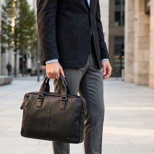 Peut inclure: Une serviette en cuir noir avec deux poignées supérieures et une bandoulière. La serviette rectangulaire a une surface texturée et des coutures visibles. La personne porte un blazer gris foncé, une chemise bleu clair et un pantalon gris.