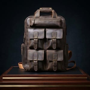 May include: A brown leather backpack with multiple front pockets secured by buckles. The backpack has a top handle, a zippered top pocket, and adjustable shoulder straps. The leather has a distressed appearance.