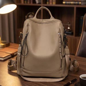 Op de afbeelding: Een beige leren rugzak met meerdere vakken met ritssluiting en verstelbare riemen. De tas heeft een handvat aan de bovenkant en een strak, modern design. De ritsen en hardware zijn donkerder van kleur, wat een contrast geeft.
