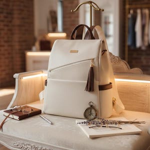 May include: Cream-colored backpack with brown accents, a tassel zipper pull, and a small gold-colored rectangular plate. The bag has a front pocket and a top handle. A compass, glasses, and a notebook are next to the bag.
