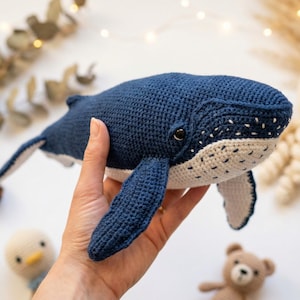 Pode incluir: Uma baleia de crochê azul marinho feita à mão com detalhes brancos na barriga e no rosto. A baleia está sendo segurada por uma mão. Outros brinquedos de crochê, incluindo um polvo, um pinguim e um urso, estão ao fundo.