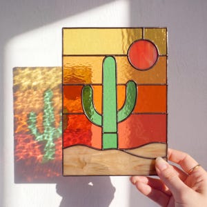 Op de afbeelding: Rechthoekig glas-in-loodpaneel met een woestijnlandschap met een groene cactus, een rode zon en een warme zonsondergang. Het paneel heeft een bruine rand. Het paneel wordt door een hand vastgehouden.