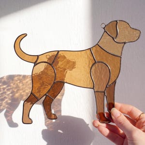 May include: A stained glass dog silhouette in shades of brown and amber. The dog is depicted in a standing pose, with a loop at the top for hanging. The artwork is held by a hand, showcasing the intricate details and color variations of the glass.