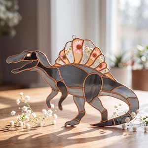 May include: A stained glass Spinosaurus dinosaur with a sail filled with pink flowers. The dinosaur is primarily gray with copper-colored outlines. The piece is hanging by a clear string, and is on a wooden surface.
