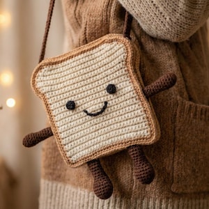 Op de afbeelding: Gehaakte crossbodytas in de vorm van een lachende toast, met armen en benen. De tas is beige met bruine details en een bruine band. De tas is ongeveer 18 cm breed en 18 cm hoog.