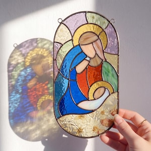 Puede incluir: Panel de vidrieras que representa una escena religiosa con la Virgen María y el niño Jesús. La obra de arte presenta una paleta de colores vibrantes que incluye azul, naranja, amarillo y verde. El panel está enmarcado con un borde de metal oscuro y tiene un lazo para colgar.