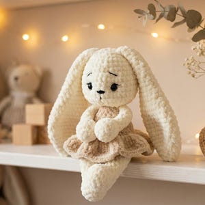 May include: A handmade, cream-colored crochet bunny doll with long, floppy ears and a tan dress. The bunny has black eyes and is sitting on a white shelf. Other toys and wooden blocks are in the background, with soft lighting.