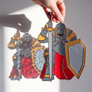 May include: A stained glass knight figure with a sword and shield. The knight is in gray, gold, and red, with a detailed design. The figure is held up by a hand, casting a shadow on the wall. The knight is a decorative item.