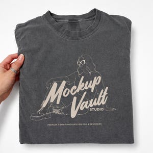 Może przedstawiać: Złożona grafitowa koszulka z grafiką osoby w okularach przeciwsłonecznych i napisem "Mockup Vault STUDIO". Koszulka ma również napis "PREMIUM T-SHIRT MOCKUPS FOR POD & DESIGNERS".
