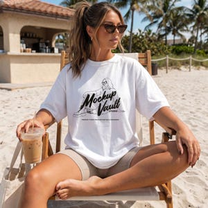 May include: White t-shirt with a black graphic of a person sitting and the text "Mockup Vault Studio". The shirt is oversized and worn with tan shorts. The person is wearing sunglasses and sitting in a wooden chair on a sandy beach.