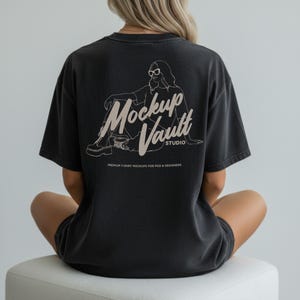 May include: Black t-shirt with a graphic on the back featuring the text "Mockup Vault Studio" and an illustration of a person wearing sunglasses. The shirt also has the text "Premium T-Shirt Mockups for POD & Designers".