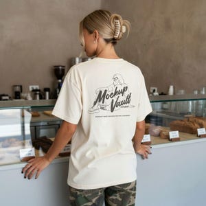 May include: Cream-colored t-shirt with a graphic on the back featuring a seated figure and the text "Mockup Vault Studio." The shirt is part of a POD (Print on Demand) design. The person is wearing camouflage pants.