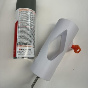 May include: A can of gray spray paint next to a white cylindrical object with a diamond-shaped cutout and an orange handle. A threaded metal rod extends from the bottom of the white object.