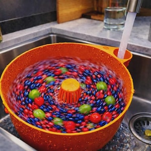 May include: An orange berry spinner filled with a mix of red strawberries, green gooseberries, and blue blueberries, being washed under a faucet. The spinner is in a stainless steel sink.