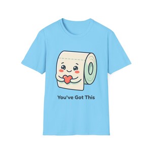 Könnte beinhalten: Hellblaues T-Shirt mit einer Cartoon-Toilettenpapierrolle, die ein rotes Herz hält. Die Toilettenpapierrolle hat ein lächelndes Gesicht und der Text "You've Got This" ist darunter aufgedruckt. Das T-Shirt ist aus weichem Material.