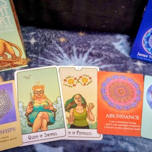 May include: A selection of tarot cards and two card deck boxes. The cards display illustrations and text, including "Queen of Swords" and "Abundance." The deck boxes are labelled "This Might Hurt" and "The Soul's Journey."