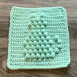May include: A light green crocheted square with a textured design in the center. The design is made up of small, raised bumps that create a 3D effect.