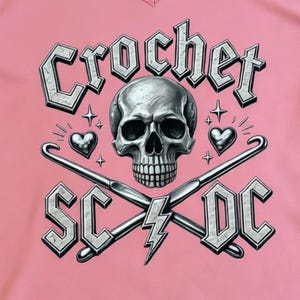 May include: A pink V-neck t-shirt featuring a skull graphic with the text "Crochet SC/DC". The design includes crossed crochet hooks, hearts, and lightning bolts in a silver color.