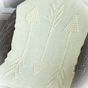 May include: Cream-colored crochet blanket featuring three arrows. The arrows are made with a textured, raised stitch, creating a 3D effect. The blanket is draped over a wicker chair. The blanket measures approximately 40 inches by 50 inches.