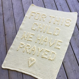 Può includere: Coperta per neonati all'uncinetto color crema. La coperta presenta il testo "FOR THIS CHILD WE HAVE PRAYED" in lettere in rilievo e strutturate. Una forma di cuore è anch'essa all'uncinetto nel design. La coperta è rettangolare e ha un bordo semplice.