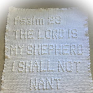 Könnte beinhalten: Weiße gehäkelte Babydecke mit dem Text "Psalm 23 Der Herr ist mein Hirte, mir wird nichts fehlen" und einem Kreuz am unteren Rand.