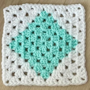 May include: A square, handmade crochet piece with a granny square design. The center is a diamond shape in a light teal color, surrounded by white. The piece is made of soft yarn and has a textured appearance, perfect for a blanket or pillow.