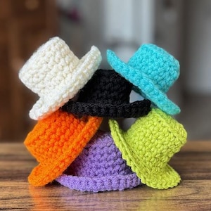 Op de afbeelding: Een stapel van zeven miniatuur gehaakte hoeden in verschillende kleuren: wit, zwart, turquoise, oranje, paars en limoengroen. De hoeden zijn gerangschikt in een piramidevorm, waarbij hun gedetailleerde haaktextuur en ontwerp worden getoond. De hoeden staan op een houten oppervlak.