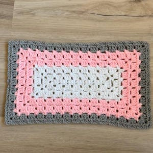 May include: A rectangular, handmade crochet rug with a granny square design. The rug features a white center, surrounded by a pink layer, and a gray border. The rug is made of soft yarn.