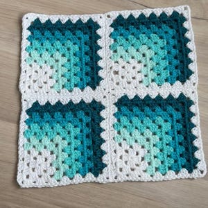 May include: A square, handmade crochet piece featuring four squares joined together. Each square has a gradient of teal and white, framed by a white border. The piece is made of yarn and rests on a wooden surface.