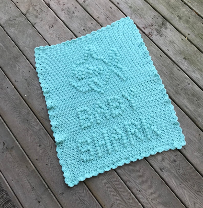 May include: A light blue crocheted baby blanket with a raised stitch pattern that spells out "Baby" in the center.