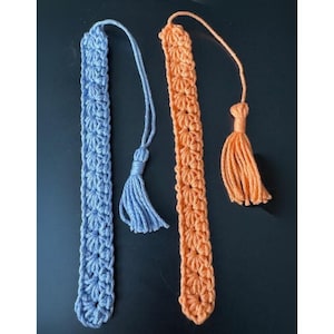 Könnte beinhalten: Zwei handgefertigte Häkel-Lesezeichen. Eines ist hellblau mit einem passenden Quasten, das andere ist orange mit einer orangefarbenen Quaste. Beide Lesezeichen haben ein Blumenmuster und sind aus Garn gefertigt.