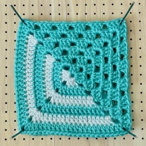 Puede incluir: Un cuadrado de ganchillo granny square turquesa con detalles blancos, sujeto a una tabla perforada de madera. El textil hecho a mano presenta un patrón calado. El diseño de ganchillo muestra la textura y la artesanía de la pieza.