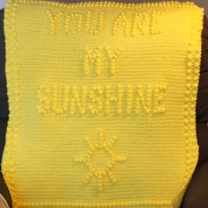 May include: Yellow crocheted blanket with the text "You are my sunshine" and a sun design in the center.